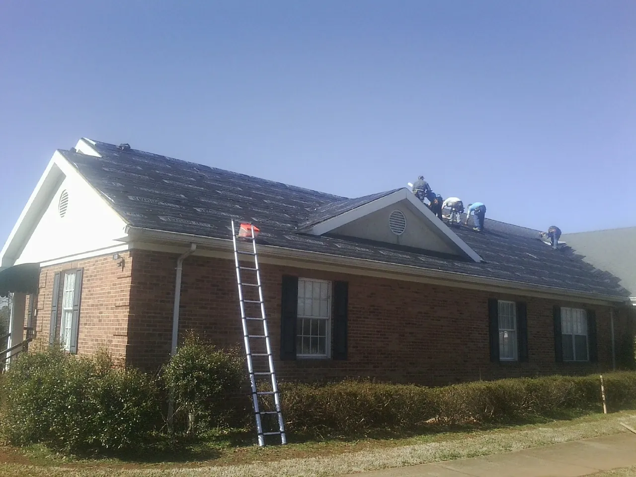Crew tearing shingles off a brick ranch roof with a ladder set — Metal Roof Repair work by our crew in Goode