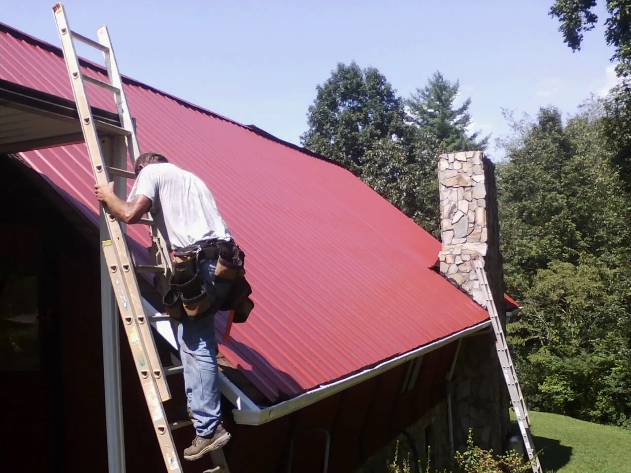 Roofer on a ladder beside a red metal roof and stone chimney — Soffit & Fascia Repair by Roofing Brothers in Goode